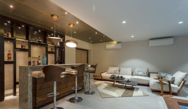 Modern home bar with stools, adjacent sitting area with sofa and chairs, warm lighting.